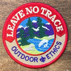 May include: A circular embroidered patch featuring a landscape with green pine trees, a blue river, and purple mountains. The red border contains the white text LEAVE NO TRACE, while the tan bottom border features the purple text OUTDOOR ETHICS.