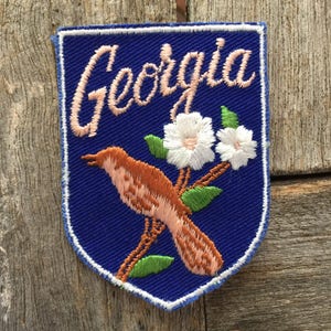 May include: Embroidered patch featuring the word "Georgia" in cursive script above a brown bird perched on a branch with white flowers and green leaves. The patch has a blue background and a white border.