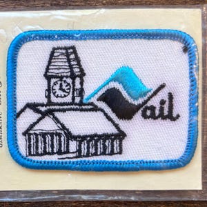 Op de afbeelding: Een blauwe en witte geborduurde patch met een zwart-witte illustratie van een gebouw met een klokkentoren en een golf. De tekst "ail" is in zwart geborduurd aan de rechterkant van de patch.