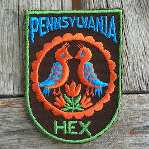 May include: A brown fabric patch with green stitching around the edges. The patch features a stylized design of two blue birds with orange and white accents, a red flower, and green leaves. The text "PENNSYLVANIA" is embroidered in blue and orange, and the text "HEX" is embroidered in green.