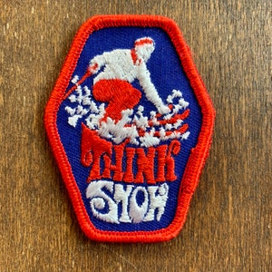 May include: A hexagonal embroidered patch with a blue background and a red border. The design features a white figure skiing on white snow with red pants. Below the skier, the text reads THINK SNOW in bold red and white embroidered lettering.