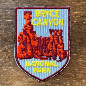May include: Embroidered patch featuring the text "BRYCE CANYON NATIONAL PARK" in yellow. The patch has a light blue background with a shield shape and a red and orange illustration of the canyon. The border is red and yellow.
