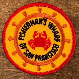 Puede incluir: Parche bordado con fondo amarillo y borde rojo. El parche presenta un cangrejo en el centro y las palabras "Fisherman's Wharf of San Francisco" en un diseño circular. Un diseño de timón de barco rodea el texto.