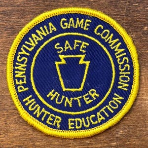 Może przedstawiać: Okrągła niebiesko-żółta naszywka z napisem "PENNSYLVANIA GAME COMMISSION" i "HUNTER EDUCATION". Na środku znajduje się żółty kształt klucza z napisem "SAFE" powyżej i "HUNTER" poniżej.