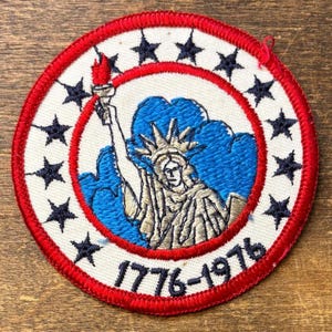 May include: Circular embroidered patch with the Statue of Liberty holding a torch, set against a blue backdrop. The design is framed by a red ring and a ring of black stars on a white background. The text "1776-1976" is at the bottom.