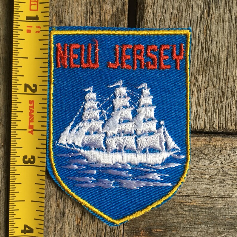 May include: A blue embroidered patch featuring the text NEW JERSEY in red letters above a white sailing ship on water. A yellow border outlines the shield shape. A yellow Stanley measuring tape shows the patch is approximately 8.9 cm tall.