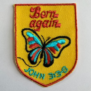 May include: A shield-shaped embroidered patch with a yellow background and a red border. It features a butterfly with turquoise and red wings, the red embroidered text "Born again," and the turquoise embroidered text "JOHN 3:3-8" at the bottom.