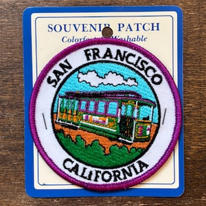 Puede incluir: Parche de recuerdo redondo con un tranvía de San Francisco. El parche tiene un borde blanco con las palabras "SAN FRANCISCO CALIFORNIA". El tranvía es verde, naranja y morado, sobre un cielo azul con nubes blancas.