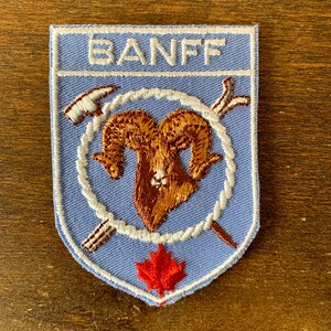 Può includere: Toppa ricamata con la scritta "BANFF" in alto. La toppa presenta una testa di ariete marrone, circondata da un cerchio di corda bianca, un piccone e attrezzi incrociati. Una foglia d'acero rossa in basso.