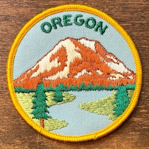 Puede incluir: Parche bordado con una escena de montaña y la palabra "OREGON" en verde. El parche tiene un fondo azul claro, una montaña naranja y blanca, árboles verdes y un borde amarillo.