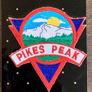 Può includere: Una toppa ricamata di colore rosso, bianco e blu con una scena di montagna e le parole "PIKES PEAK" su uno stendardo bianco.