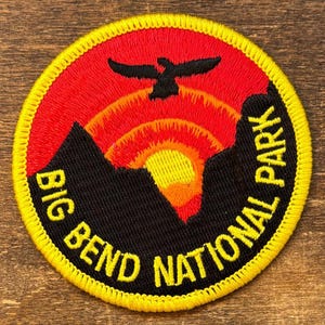 May include: Circular embroidered patch featuring the text "BIG BEND NATIONAL PARK" in yellow on a black background. The patch has a red and orange sunset design with a black mountain silhouette and a black bird.