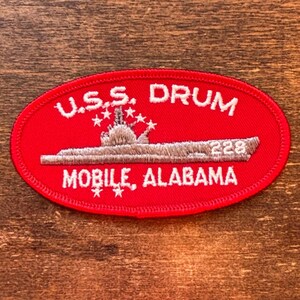 May include: Oval red patch with white embroidered text and a submarine illustration. The text reads "U.S.S. DRUM MOBILE, ALABAMA 228". The patch features a detailed depiction of a submarine and stars.