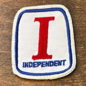 May include: A rectangular embroidered patch with rounded corners featuring a white background, a blue border, and a white outer edge. A large red letter I is centred above the blue text INDEPENDENT, which is stitched in a bold, sans-serif font.