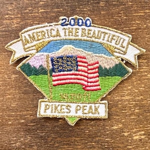 Può includere: Toppa ricamata con la bandiera americana davanti a una catena montuosa. La toppa recita "America the Beautiful" e "Pikes Peak 2000" in lettere dorate e bianche. La toppa ha uno schema di colori verde, blu e rosa.