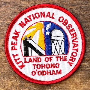 May include: A circular embroidered patch with a red border and white background. It features a yellow sun, a black and white structure, and a blue observatory dome. Text reads: KITT PEAK NATIONAL OBSERVATORY LAND OF THE TOHONO O'ODHAM.