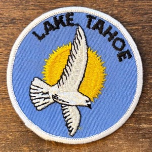 Könnte beinhalten: Gestickter Aufnäher mit einem weißen Vogel, der vor einer gelben Sonne auf hellblauem Hintergrund fliegt. Die Worte "LAKE TAHOE" sind in Schwarz um den Rand gestickt. Der Aufnäher hat einen weißen Rand.
