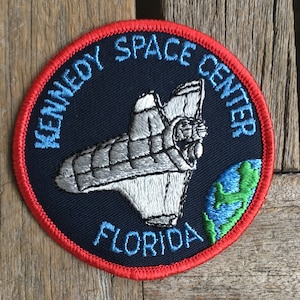 Może przedstawiać: Okrągła naszywka z haftem z czerwoną obwódką, przedstawiająca logo Kennedy Space Center. Środek w kolorze granatowym przedstawia srebrny prom kosmiczny, Ziemię i napis "KENNEDY SPACE CENTER FLORIDA".