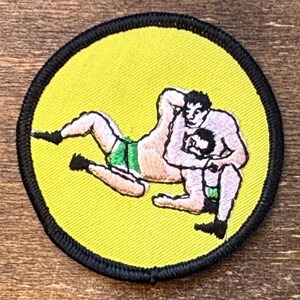 Könnte beinhalten: Runder Aufnäher mit einer Wrestling-Szene. Zwei Ringer sind im Kampf, einer trägt grüne Shorts. Der Aufnäher hat einen gelben Hintergrund und einen schwarzen Rand.