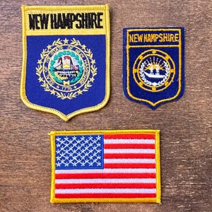 May include: Three embroidered patches: two with "NEW HAMPSHIRE" and state seal designs, and one American flag. The patches are blue, gold, red, and white, with detailed stitching. The patches are likely for clothing or accessories.