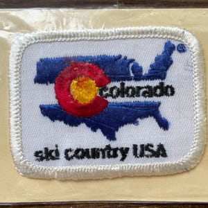 Op de afbeelding: Geborduurde patch met de omtrek van Colorado in blauw, met een rood en geel cirkelvormig embleem. Het woord "colorado" staat in zwarte tekst. De zin "ski country USA" staat eronder in zwarte tekst, op een witte achtergrond.