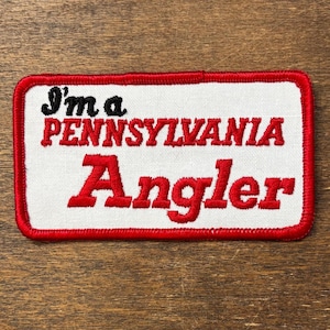 Puede incluir: Parche bordado con el texto "I'm a PENNSYLVANIA Angler" en letras negras y rojas. El parche tiene un fondo blanco con un borde rojo. El parche es de forma rectangular.