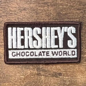 May include: A rectangular embroidered patch with a dark brown background and a stitched border. The text HERSHEY'S is embroidered in large white capital letters, with the words CHOCOLATE WORLD in smaller white capital letters underneath.