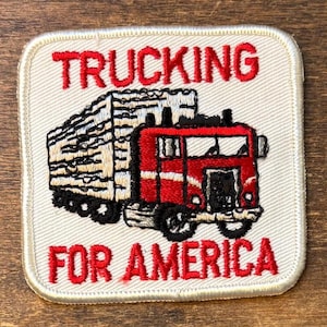May include: Embroidered patch with a red and black semi-truck hauling a load. The patch has the words "TRUCKING" above the truck and "FOR AMERICA" below in red, all on a white background with a stitched border.