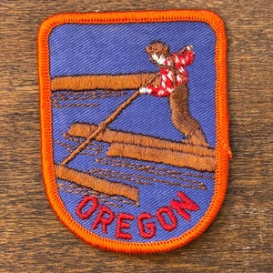 Puede incluir: Parche bordado con borde naranja y fondo azul. El parche presenta una figura de leñador con camisa a cuadros rojos y blancos, equilibrándose sobre troncos. La palabra "OREGON" está cosida en rojo en la parte inferior.