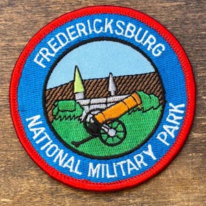 Op de afbeelding: Ronde patch met een rode rand en blauwe achtergrond. De patch heeft de tekst "FREDERICKSBURG NATIONAL MILITARY PARK" in het wit. Het midden toont een kanon, gebouwen en groen gras onder een blauwe lucht.