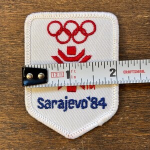 Puede incluir: Parche blanco con anillos ol&iacute;mpicos y emblema rojos, y el texto "Sarajevo '84" en azul. El parche mide aproximadamente 5 cm de ancho. El parche est&aacute; sobre una superficie de madera.
