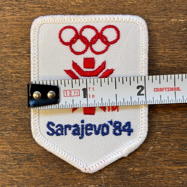 Puede incluir: Parche blanco con anillos ol&iacute;mpicos y emblema rojos, y el texto "Sarajevo '84" en azul. El parche mide aproximadamente 5 cm de ancho. El parche est&aacute; sobre una superficie de madera.