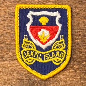 May include: A shield-shaped embroidered patch with a dark blue background and a yellow stitched border. The center features a white-bordered crest with a red field, a yellow shell icon, and a yellow plant graphic. A yellow banner reads JEKYLL ISLAND.