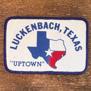 May include: A rectangular patch with a white background and a blue border. The patch features a blue and red outline of the state of Texas with a white star. The text "LUCKENBACH, TEXAS" arches above the state outline, and "UPTOWN" is written below.