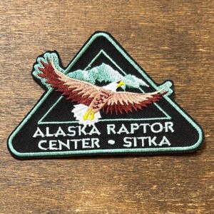 May include: Embroidered patch featuring a soaring eagle with outstretched wings, set against a mountain backdrop within a black triangle. The text "ALASKA RAPTOR CENTER • SITKA" is displayed in white.