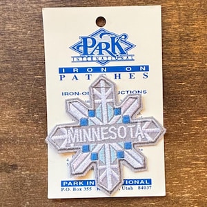 May include: A grey embroidered iron-on patch in the shape of a snowflake, with the word "MINNESOTA" in the centre. The patch has blue and light blue accents and is attached to a white card with the "PARK INTERNATIONAL" logo.