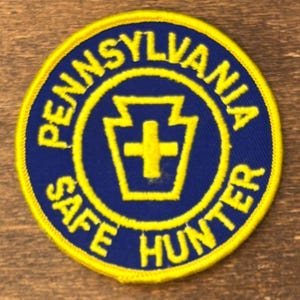 Może przedstawiać: Okrągła naszywka z niebieskim tłem i żółtym tekstem i akcentami. Tekst brzmi "PENNSYLVANIA SAFE HUNTER" na obwodzie. Żółty kamień węgielny z krzyżem znajduje się na środku.