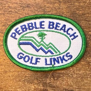 Puede incluir: Un parche bordado de forma ovalada con fondo blanco y borde verde. El texto PEBBLE BEACH GOLF LINKS está bordado en azul. Un gráfico central muestra la silueta de un árbol verde sobre líneas onduladas de color azul y verde.