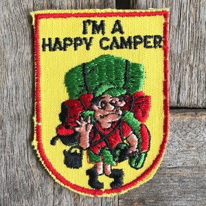 May include: Yellow embroidered patch with red trim featuring a cartoon character wearing a green and red backpack and the text "I'm a Happy Camper".