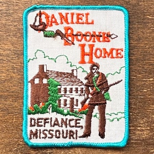May include: Embroidered patch with a teal border, featuring the text "Daniel Boone Home, Defiance, Missouri." The patch depicts a brown house, a figure with a rifle, and a bow and arrow.