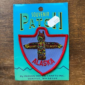 以下が含まれることがあります： カラフルなトーテムポールデザインと赤い刺繍の「ALASKA」の文字が特徴の、お土産パッチ。パッチは赤い縁取りのある青色の背景にあり、上部には「SOUVENIR PATCH」の文字があります。