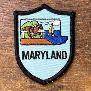 May include: A shield-shaped embroidered patch with a light blue background and a black border. The design features a brown horse with a yellow rider, a white fence, and a sailboat on blue water. The word MARYLAND is stitched in black letters.