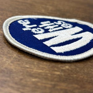 Puede incluir: Un parche bordado circular con fondo azul oscuro y borde blanco. El parche presenta letras grandes y llamativas bordadas en blanco que forman la palabra HI, con un texto bordado en blanco m&aacute;s peque&ntilde;o situado sobre la letra H.