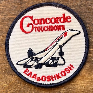 May include: A white ceramic mug with a black handle and the text "But First, Coffee" in a bold, black font. The mug is set against a blurred background. The mug appears to be approximately 10 cm tall.