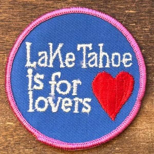 Könnte beinhalten: Ein runder Aufnäher mit blauem Hintergrund und dem weißen Text "Lake Tahoe is for lovers". Auf der rechten Seite befindet sich ein rotes Herz. Der Aufnäher hat einen rosa genähten Rand.