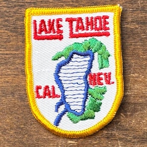 Könnte beinhalten: Gestickter Aufnäher mit "LAKE TAHOE" in roten Blockbuchstaben. Der Aufnäher zeigt einen blauen Seeumriss mit weißen Linien, grüne Bäume und die Buchstaben "CAL" und "NEV". Der Aufnäher hat einen gelben Rand.