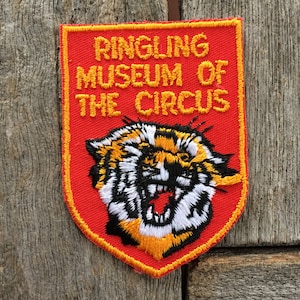 Könnte beinhalten: Ein schildförmiger, bestickter Aufnäher mit rotem Hintergrund und gelbem Rand. Der Text RINGLING MUSEUM OF THE CIRCUS ist in gelbem Garn gestickt. Unter dem Text befindet sich ein detaillierter, gestickter Tigerkopf in Orange, Weiß und Schwarz.