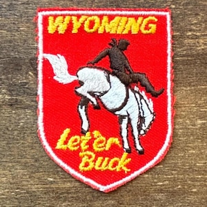 May include: A shield-shaped embroidered patch with a red background and a white border. It features the yellow text WYOMING at the top and Let 'er Buck at the bottom. A dark brown figure rides a white bucking horse in the center of the patch.