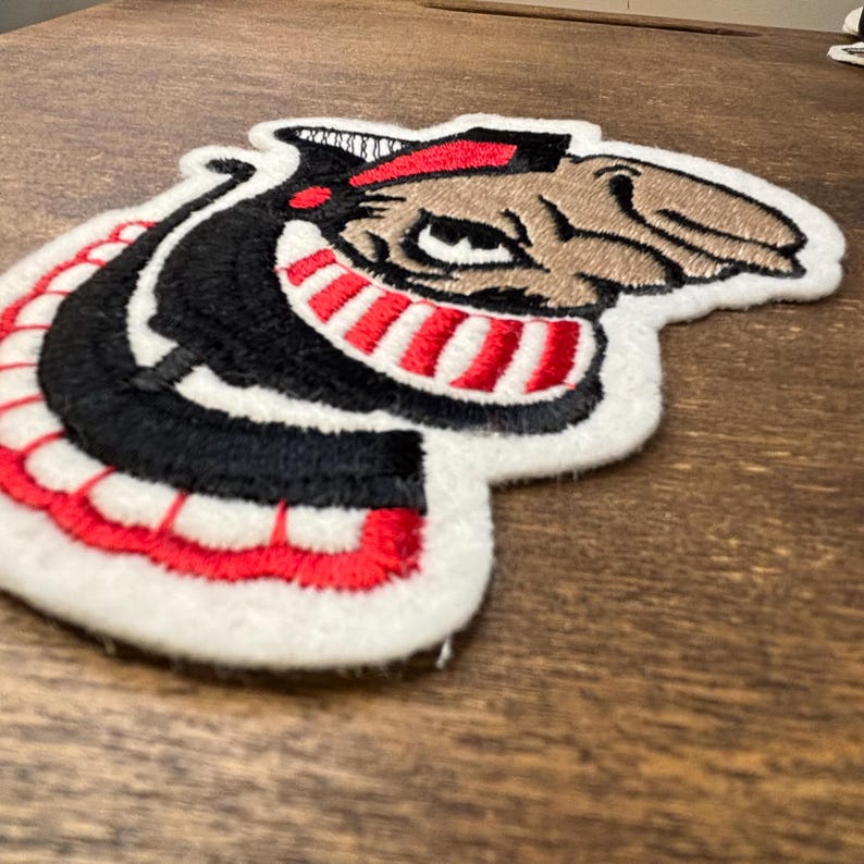 Pu&ograve; includere: Una toppa ricamata raffigurante una testa di mascotte stilizzata in filo nero, bianco, rosso e marrone chiaro. Il design mostra un personaggio con un naso prominente, un ampio sorriso a strisce rosse e bianche e un berretto nero, bordato in feltro bianco.