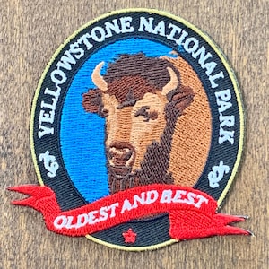 May include: A circular embroidered patch featuring a brown bison head centered on a blue background. The black border reads Yellowstone National Park in white text. A red ribbon banner at the bottom displays the white text Oldest and Best.